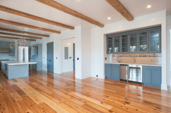 Modern kitchen with wooden floors, exposed wooden ceiling beams, blue-gray cabinets, glass cabinet doors, a built-in wine fridge, a large island, and white walls with recessed and pendant lighting.
